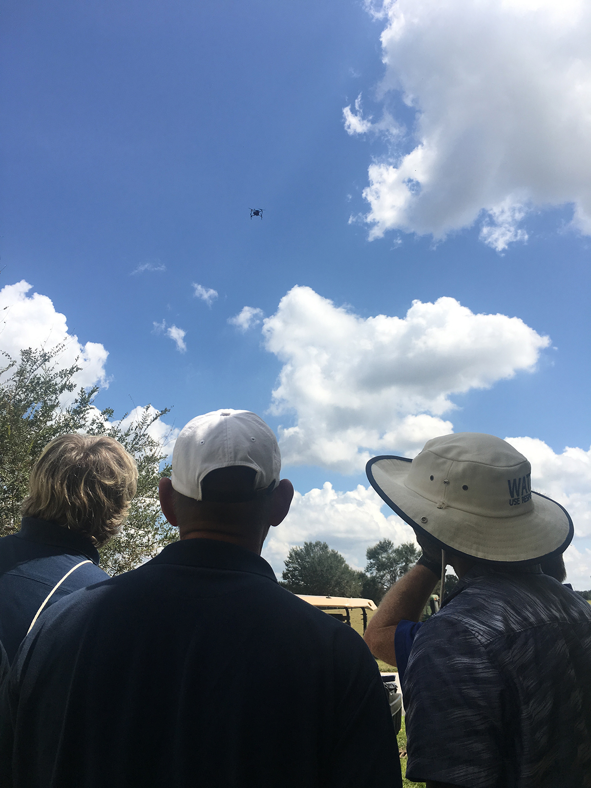 Photo of people looking up, watching a drone flying in a blue sky.